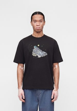 Mand med vitiligo iført sort t-shirt med space-tema sneakers grafik og blå jeans, stående mod en ensfarvet hvid baggrund.