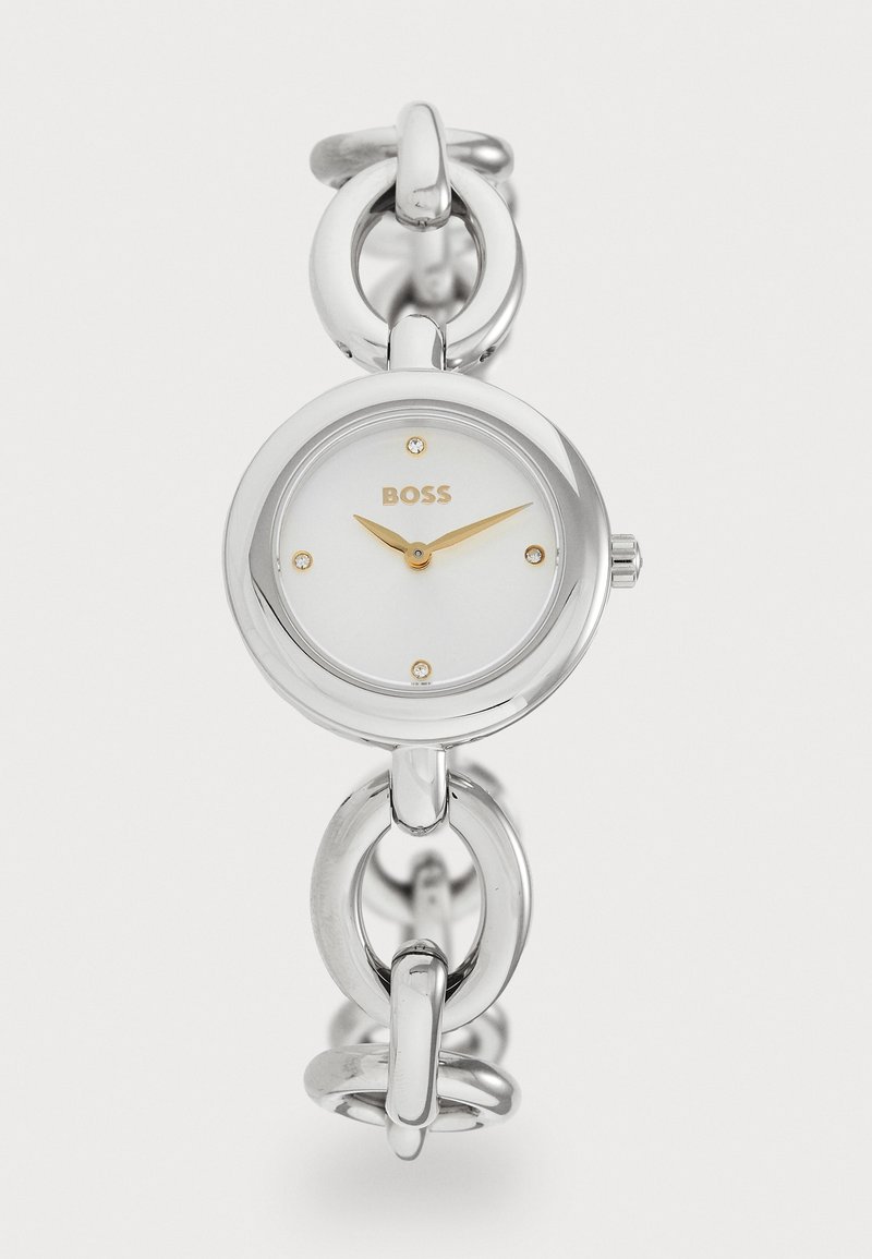 Ceas de mână din metal argintiu cu un cadran rotund, față albă, ace de oră aurii și patru accente de cristale. Design cu brățară din lanț.