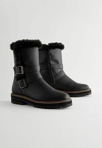 Mustad mustad mustad valge faux karvaga, kaks reguleeritavat rihma pandlatega ja paks tumbi tallaga. Omab küljeavet hõlpsaks selga panemiseks.