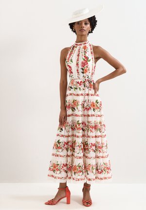 Femme portant une robe blanche à col halter avec des motifs floraux rouges et roses, un chapeau blanc à large bord et des sandales à talons hauts couleur corail.