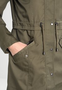 Olivgrön jacka med justerbar midja, dragkedjefickor på sidan och tryckknappar. Tillverkad av hållbart material med en slät yta.