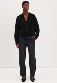 Schwarze, flauschige Jacke mit Reißverschluss und hohem Kragen, kombiniert mit hochgeschnittenen, dunklen Jeans und spitzen, schwarzen Schuhen mit silbernen Nieten.