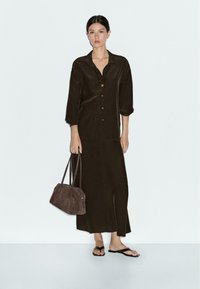 Woman wearing dark brown long skirt and button-up shirt, holding brown handbag, wearing black sandals, standing against plain light background.