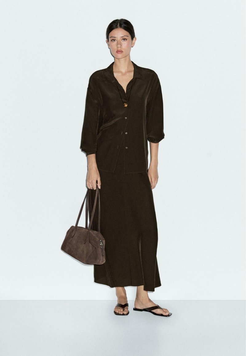 Woman wearing dark brown long skirt and button-up shirt, holding brown handbag, wearing black sandals, standing against plain light background.