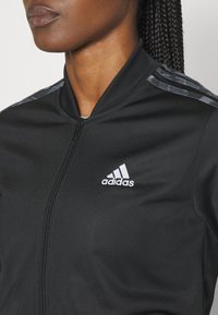 Čierna zipová bunda vyrobená z hladkého materiálu, s rebrovaným golierom, tromi šed�ými pruhmi na pleciach a bielym logom Adidas.