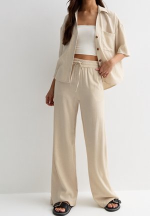 Žena ve světle hnědých volných kalhotách se stahovací šňůrkou, sladěné krátké košili na knoflíky, bílém topu bez ramínek a černých sandálech s přezkami.