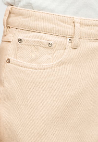 Κοντινή λήψη μπεζ τζιν παντελονιού που δείχνει την μπροστινή τσέπη με καρφίτσες, θηλιά ζώνης και διακριτικό κεντητό λογότυπο στην επένδυση της τσέπης.