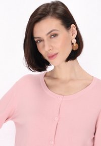 Cárdigan rosa de botón con un cuello redondo y mangas cortas. El modelo lleva pendientes redondos que presentan una mezcla de tonos beige y marrón.