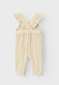 Beige baby romper med korslagda volangaxelremmar och elastisk midja, tillverkad av lätt strukturerad tyg.