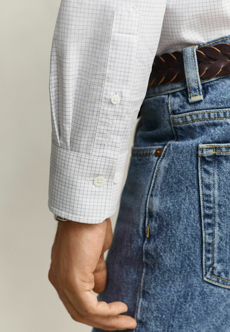 Rechter Arm einer Person mit weiß kariertem Hemd und blauen Jeans mit einem braunen, geflochtenen Gürtel.