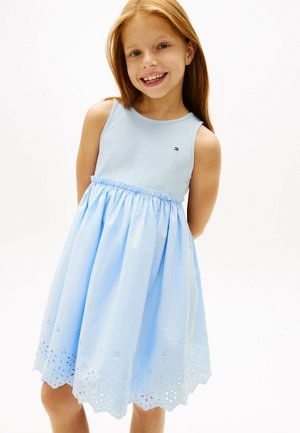 Joven sonriente con cabello rojo largo, vestida con un vestido azul claro sin mangas con detalles florales calados en el dobladillo, de pie sobre un fondo blanco.