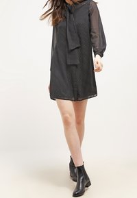 Abito nero a pois con collo ad annodare, maniche lunghe trasparenti e vestibilità ampia. Abbinato a stivaletti neri, con una texture liscia.