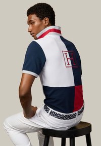 Homme assis sur un tabouret portant un polo à blocs de couleurs bleu marine, blanc et rouge avec un col relevé, ainsi qu'un pantalon blanc avec une ceinture à motif.