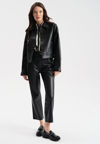 Giacca cropped in pelle nera con bottoni a pressione, abbinata a pantaloni a vita alta dello stesso colore e mocassini neri lucidi. Camicia a righe sotto.