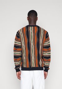 Strickpullover in Orange, Schwarz und Grün mit wellenförmigen Streifenmustern, gerippten Bündchen und Saum. Weiche Textur mit grafischen Details.