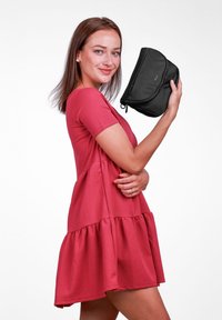 Černá psaníčko držené v ruce, má hladkou texturu, zaoblený tvar a zipové uzavírání. Modelka má na sobě růžové, vrstvené šaty.