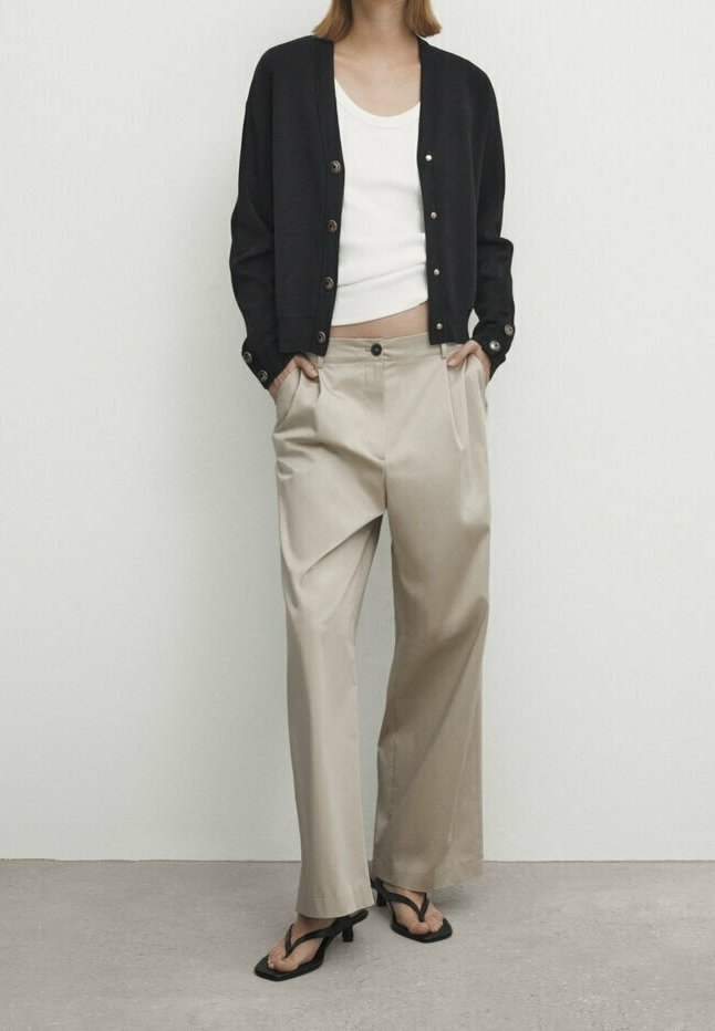 Czarny kardigan z złotymi guzikami, biały top na ramiączkach, beżowe szerokie spodnie i czarne klapki na jasnoszarym tle.