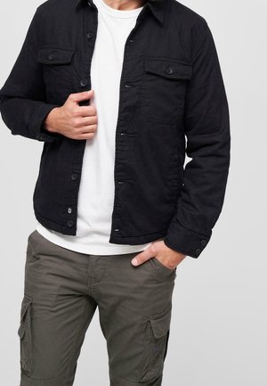 Homme portant une veste noire boutonnée sur une chemise blanche avec un pantalon cargo vert olive, une main dans la poche, l’autre tenant la veste ouverte.