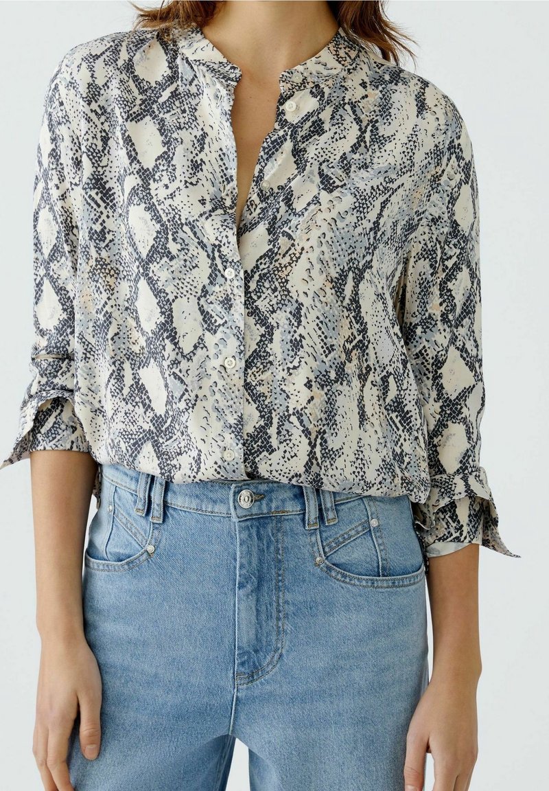 Femme portant un chemisier à manches longues imprimé serpent rentrée dans un jean en denim bleu clair taille haute, sur un fond uni.