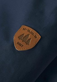 Tejido azul marino con un acabado texturizado, que presenta un parche de cuero marrón grabado con árboles y coordenadas, sujeto con bordes cosidos.