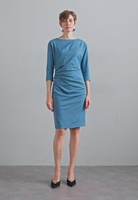 Vestido azul a la altura de la rodilla con mangas tres cuartos, frente drapeado y silueta ajustada, combinado con tacones negros de punta.