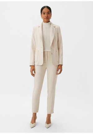 Costume beige clair ajusté avec une veste cintrée dotée de deux poches avant, associé à un pantalon slim et des talons aiguilles assortis.