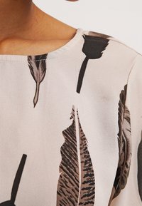Top beige chiaro con scollo rotondo e motivo di piume in toni di nero e marrone, con texture del tessuto liscia e vestibilità comoda.