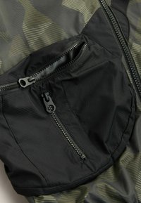 Tasca nera con zip su un tessuto leggero con un motivo camouflage in diverse tonalità di verde e nero, caratterizzata da una superficie liscia e una cucitura dettagliata.