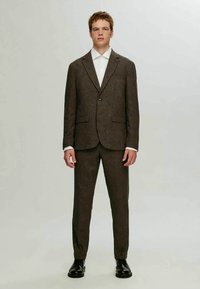 Fato castanho texturizado com reverso em ponta, blazer de botão único, calças a condizer e uma camisa branca por baixo. Sapatos pretos completam o look.