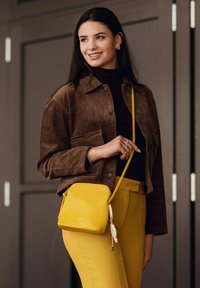 Žlutá kožená kabelka přes rameno s texturovaným povrchem, vybavená zipovým uzávěrem a ozdobným střapcem. Kombinováno s hnědým sametovým sakem a žlutými kalhotami.