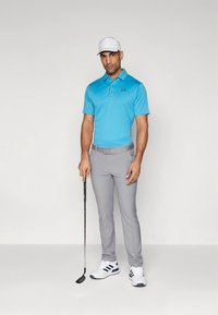 Polo azul claro com gola, calças cinzentas e sapatos brancos com detalhes azuis, segurando um taco de golfe, a usar um boné branco.