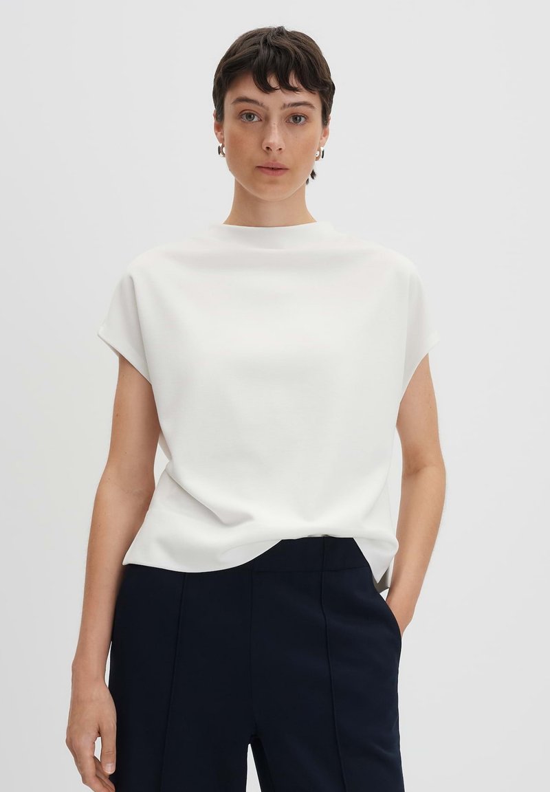 Haut blanc à manches courtes avec un col montant et une coupe décontractée, associé à un pantalon sur mesure bleu marine, avec une texture lisse.