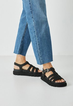 Black braided sandals with a chunky sole, featuring multiple straps and a metallic buckle, worn with cropped blue denim jeans.