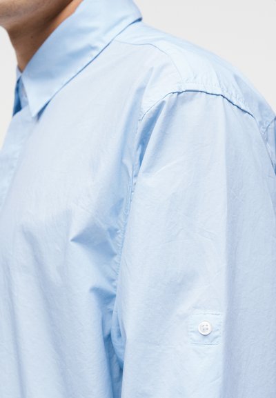 Ανοιχτό μπλε πουκάμισο με μακριά μανίκια και λεπτομέρεια με κουμπί στο μανίκι, φορεμένο από άτομο με μερικώς ορατό λαιμό και ώμο.