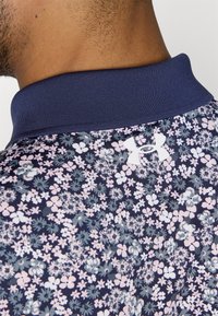 Marinblå polo med blommönster i rosa och ljusblått. Har en ribbad krage och en vit logotyp på baksidan. Mjuk tygkänsla.