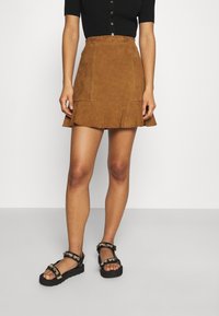 Mini-jupe en daim marron avec un ourlet évasé et des poches avant. Associée à des sandales noires ornées de bijoux dorés décoratifs.