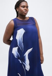 Γυναίκα που φοράει αμάνικο φόρεμα σε σκούρο μπλε χρώμα με διαφανές επάνω μέρος και μεγάλο λευκό φλοράλ μοτίβο μπροστά, στέκεται μπροστά σε φωτεινό φόντο.