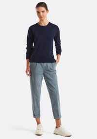 Mujer de pie con las manos en los bolsillos, vistiendo un suéter de manga larga azul marino, pantalones claros de tiro alto y zapatillas blancas sobre fondo blanco.