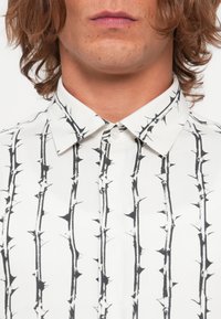 Hvit skjorte med et svart tornmønster og knapp-ner-kant. Stoffet har en glatt tekstur og er lett.