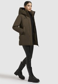 Olive green puffer jacket with a large hood, zip closure, and side pockets. Worn over a black top and paired with black faux leather leggings and boots.