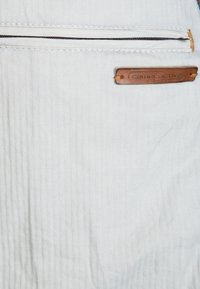 Gros plan d'un tissu beige clair avec une poche zippée horizontale et une petite étiquette en cuir marron portant la mention "camel active."