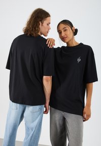 Svart oversized t-skjorter laget av bomull, med en liten hvit slange-logo på brystet, parret med lyse og mørke denimjeans.