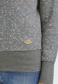 Sudadera gris con un patrón disperso de pequeños puntos rosas, azules y blancos. Presenta puños y dobladillo acanalados, con una pequeña etiqueta de marca.