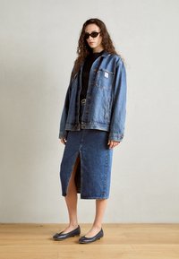 Chaqueta de denim con bolsillos frontales, ajuste oversize; suéter negro de canalé debajo; falda de denim con alta abertura; planos grises con detalle de lazo.