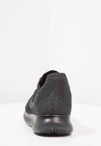 Chaussure de sport noire avec une tige en maille texturée, un talon arrondi et une semelle lisse et flexible avec des motifs de rainures distincts.