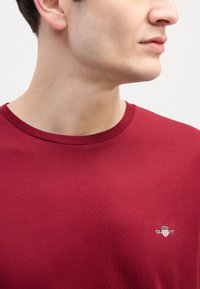 Maroon crew-neck t-paita, joka on valmistettu pehmeästä puuvillasta ja jossa on pieni valkoinen logo vasemmassa rinnassa. Tasainen rakenne ja klassinen istuvuus.