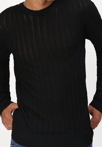 Maglione nero lavorato a maglia con un motivo a coste, maniche lunghe e scollatura a girocollo. Il tessuto ha una consistenza leggermente trasparente con strisce verticali.