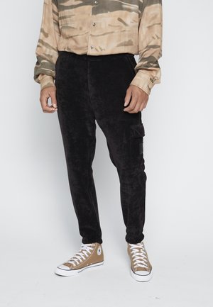 Person trägt schwarze Cord-Cargohosen, beige-schwarzes Camouflage-Knopfshemd und beige High-Top-Sneaker mit weißen Schnürsenkeln.