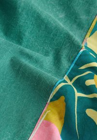 Gros plan sur un tissu bleu sarcelle avec une couture reliant un matériau vert uni à une partie à motifs avec des formes abstraites jaunes, roses et crème.