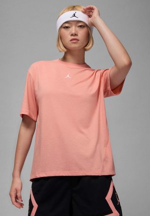 Rosa bomull T-skjorte med rund hals, korte ermer og en hvit Jordan-logo på brystet. Modellen har på seg et hvitt hårbånd.